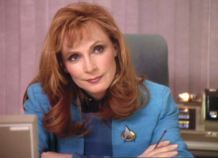 Doctor Beverly Crusher in a blue Starfleet medical uniform, leaning forward at a workstation.