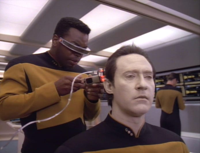 Geordi La Forge working behind Data's head with a diagnostic device aboard the Enterprise-D.