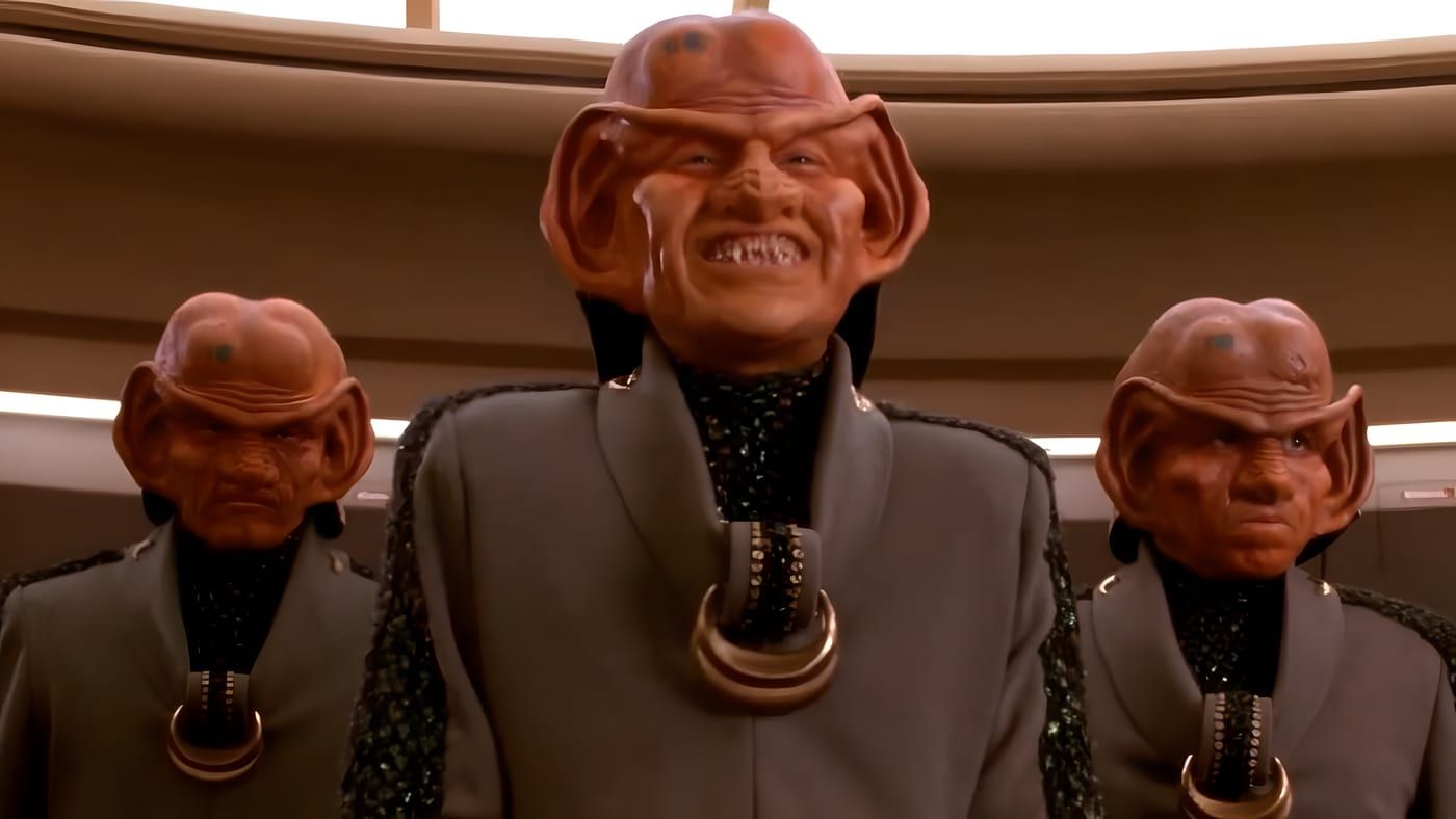 A Ferengi male in ornate robes and jewelry, standing in a dim interior.