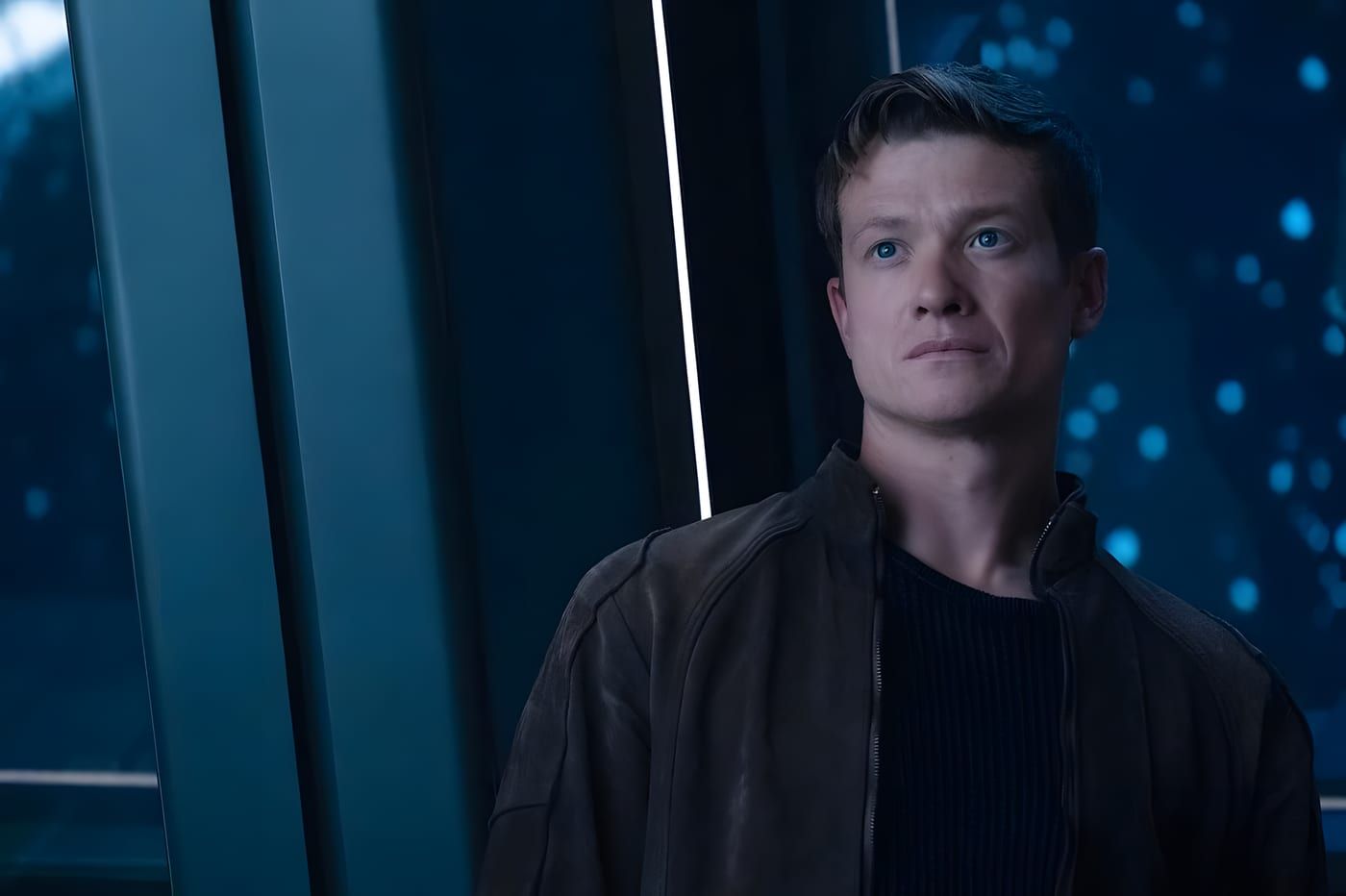 Jack Crusher in a dark jacket over a charcoal shirt, serious expression, starship corridor with a vertical light strip behind him.