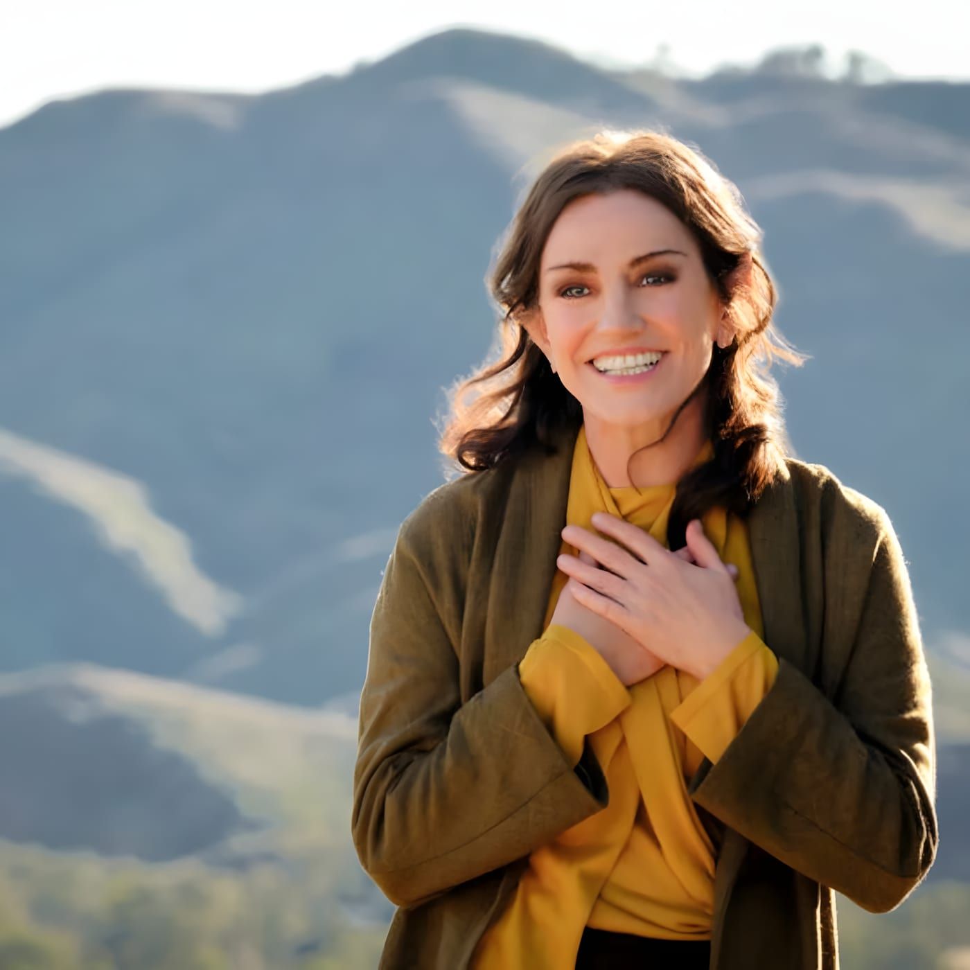 Laris outdoors with hills behind her, smiling with both hands over her heart, wearing layered mustard and olive clothing.