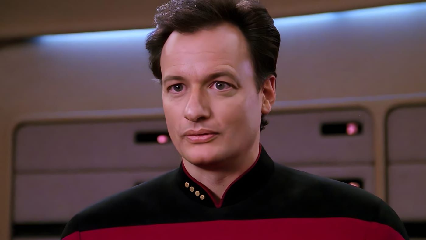 Q in a Starfleet captain's uniform with red shoulders, standing on the Enterprise-D bridge.
