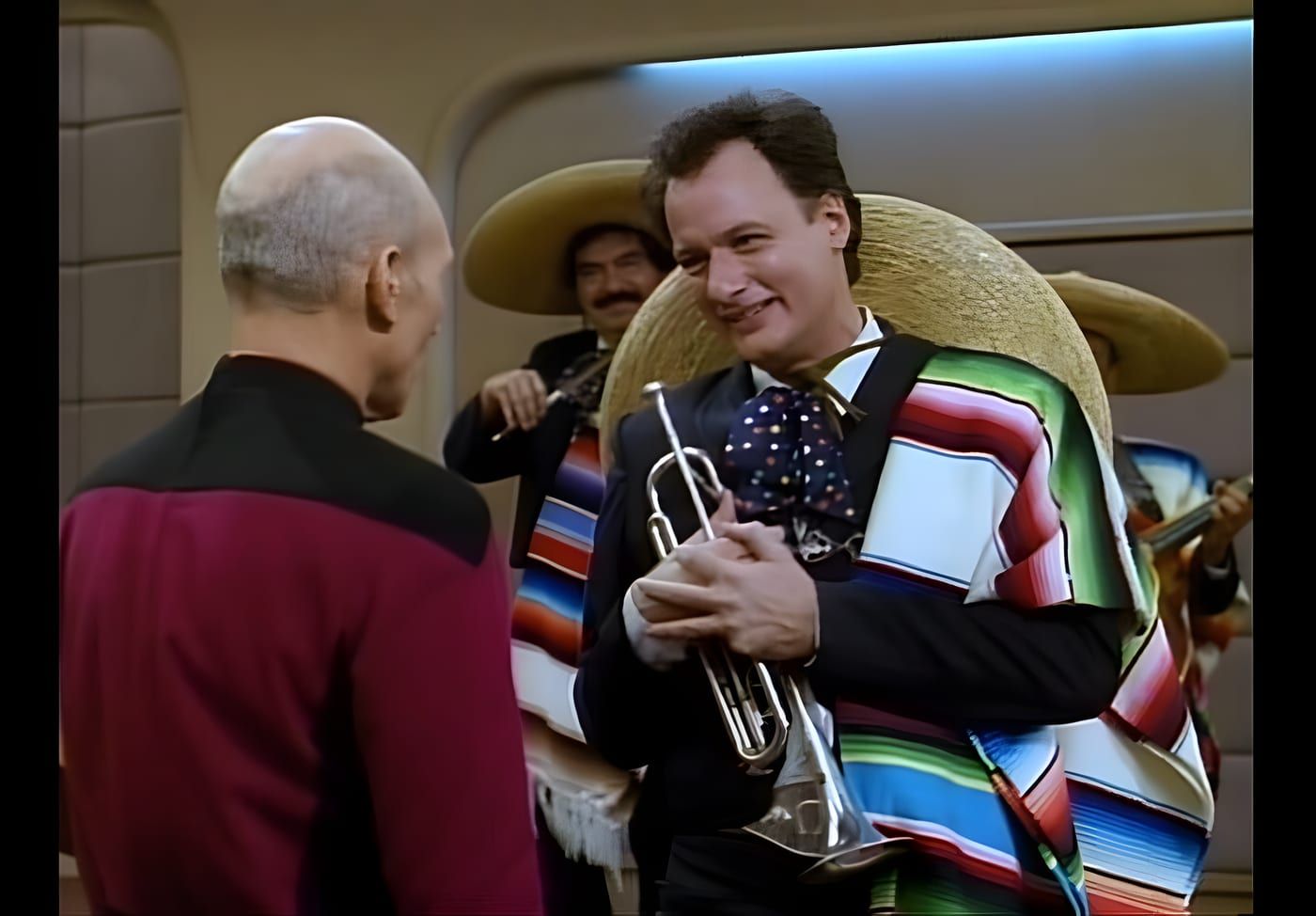 Q in mariachi dress with a trumpet, a mariachi band behind him, facing Picard on the Enterprise-D bridge.