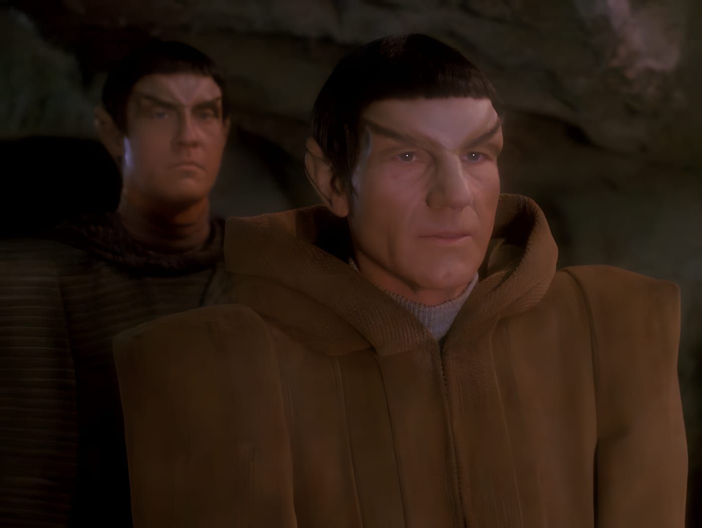 Picard in Romulan disguise with hooded brown robes and Romulan brow, another Romulan figure behind him in a dim rocky cavern.