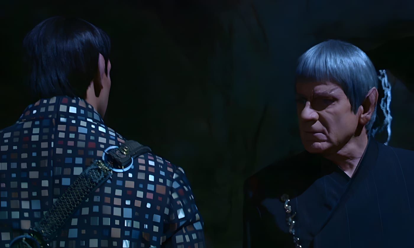 Ambassador Spock in Romulan civilian robes facing Picard, who is seen from behind in a dark mosaic-pattern jacket, in a cavern on Romulus.