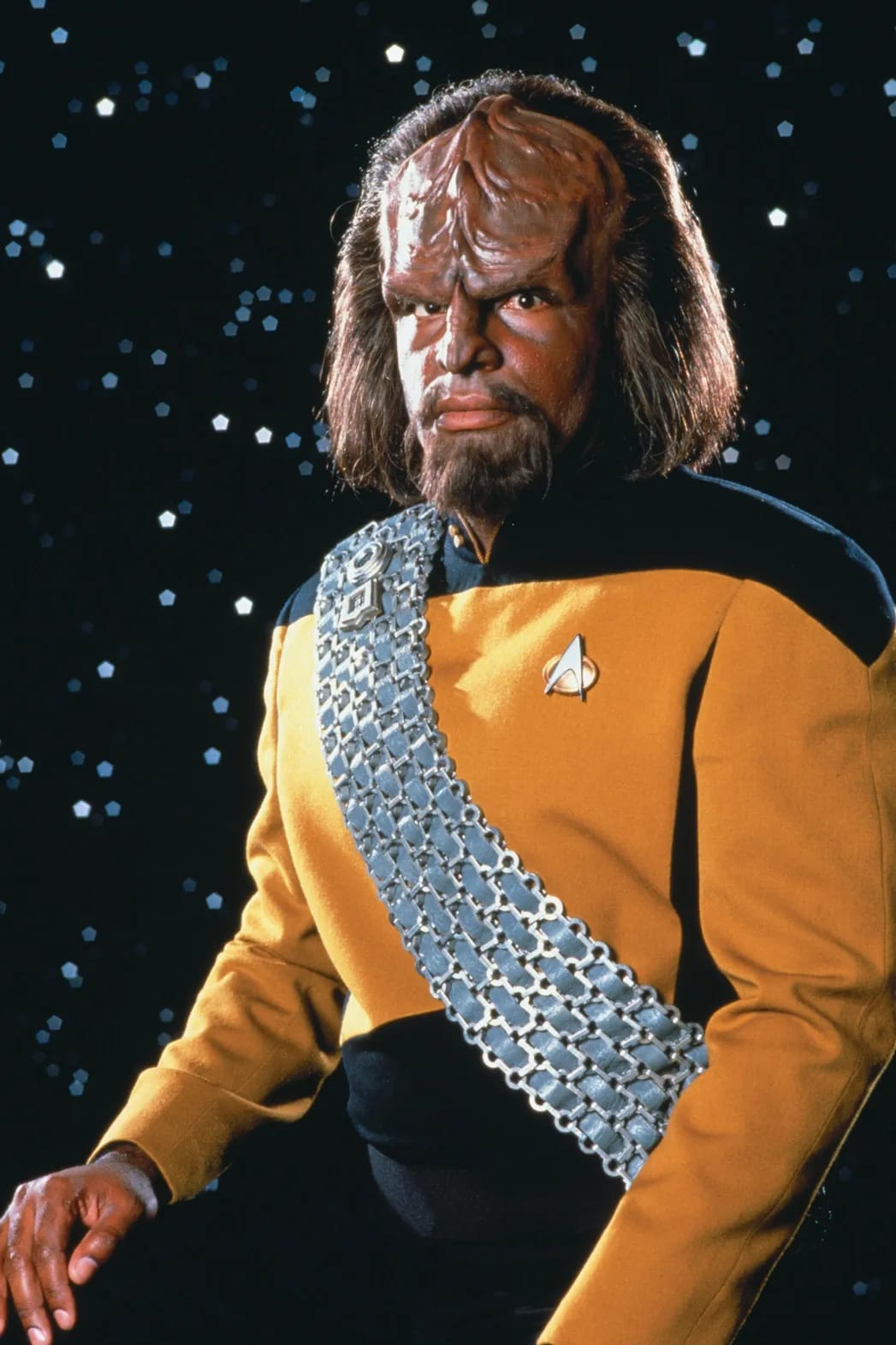 Worf standing in a gold Starfleet uniform and Klingon baldric.
